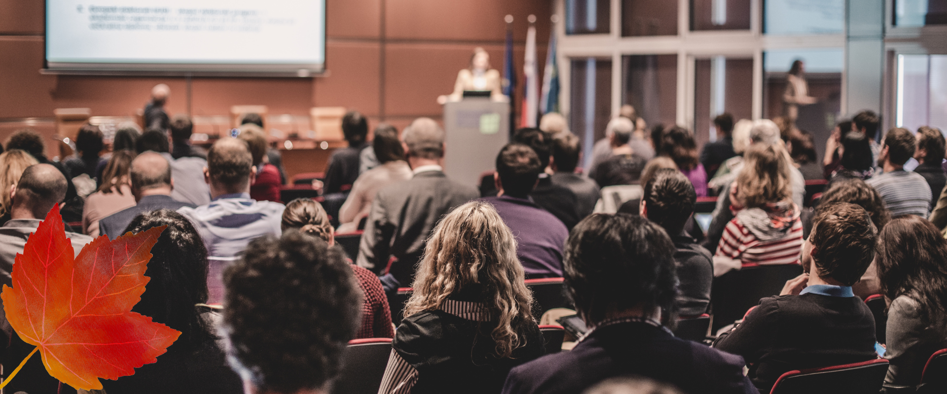 Audience seated in a conference hall facing a speaker at a podium; a large orange autumn leaf is in the lower left foreground.